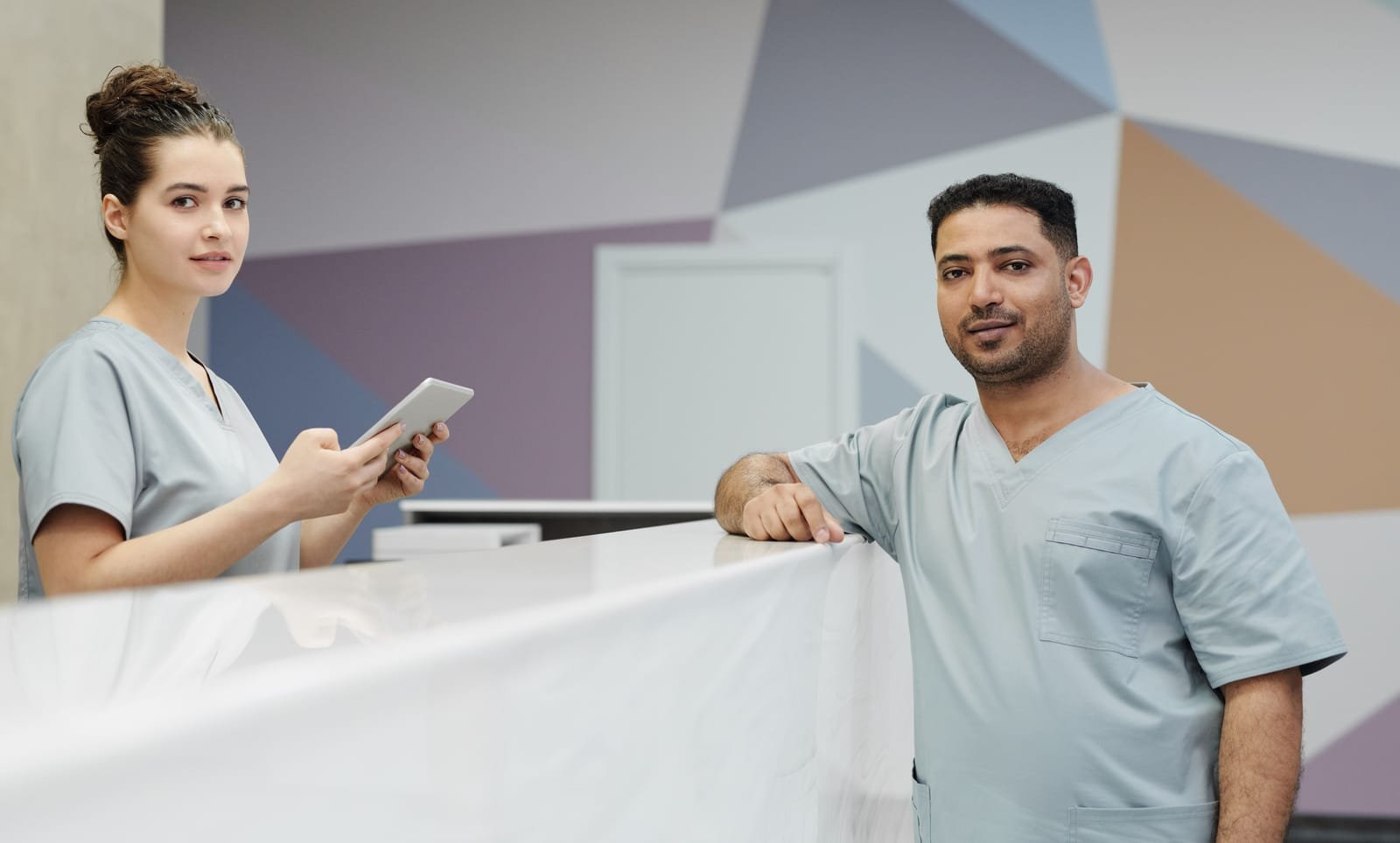 Two staff members in scrubs at the reception counter of a clinic, one holding a tablet — the entrance area a security and safety assessment starts with. Photo: Cedric Fauntleroy, via Pexels (Pexels License)