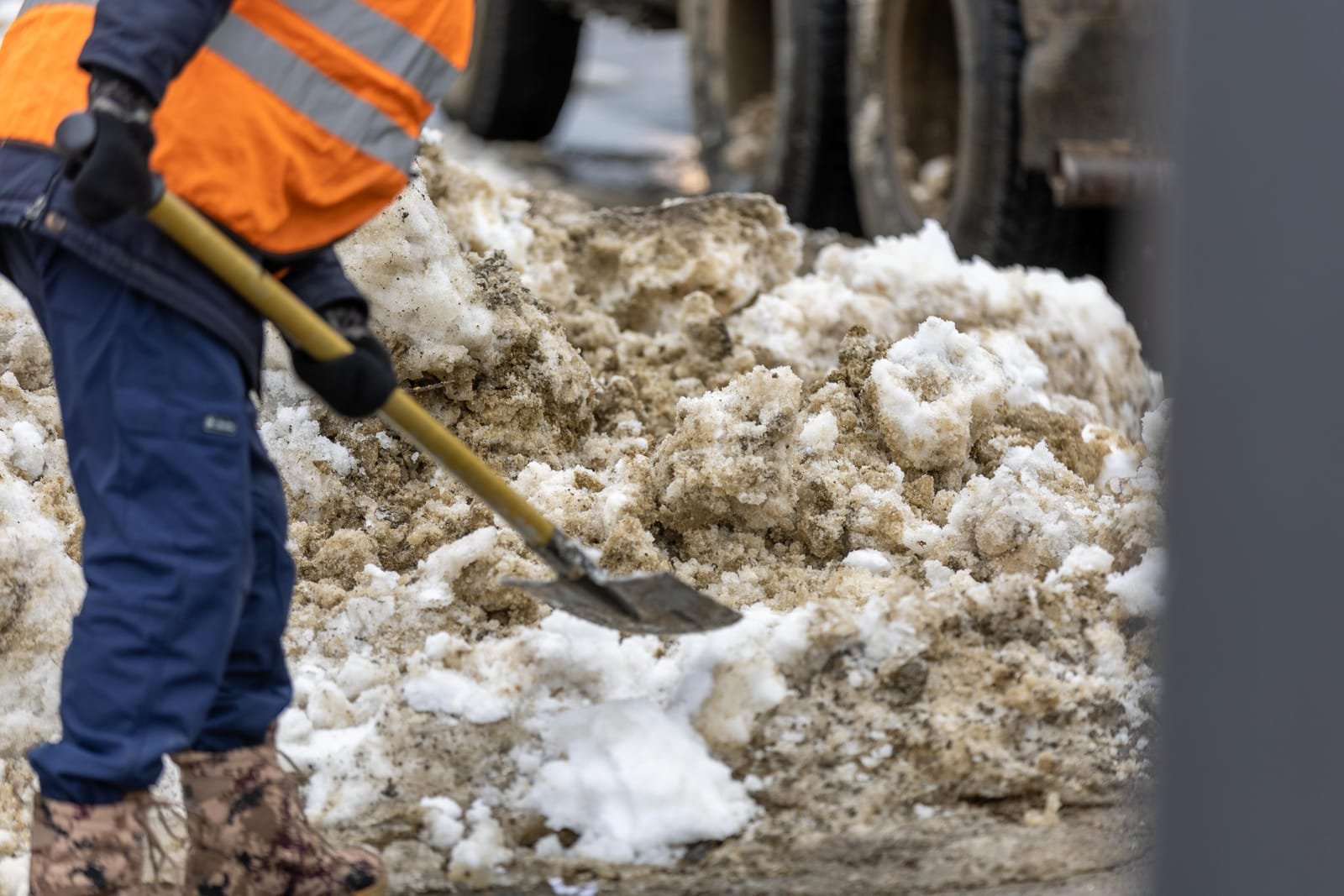A worker in a high-visibility jacket and insulated trousers shovels packed snow and slush beside a truck. Photo: Sergei Starostin, via Pexels