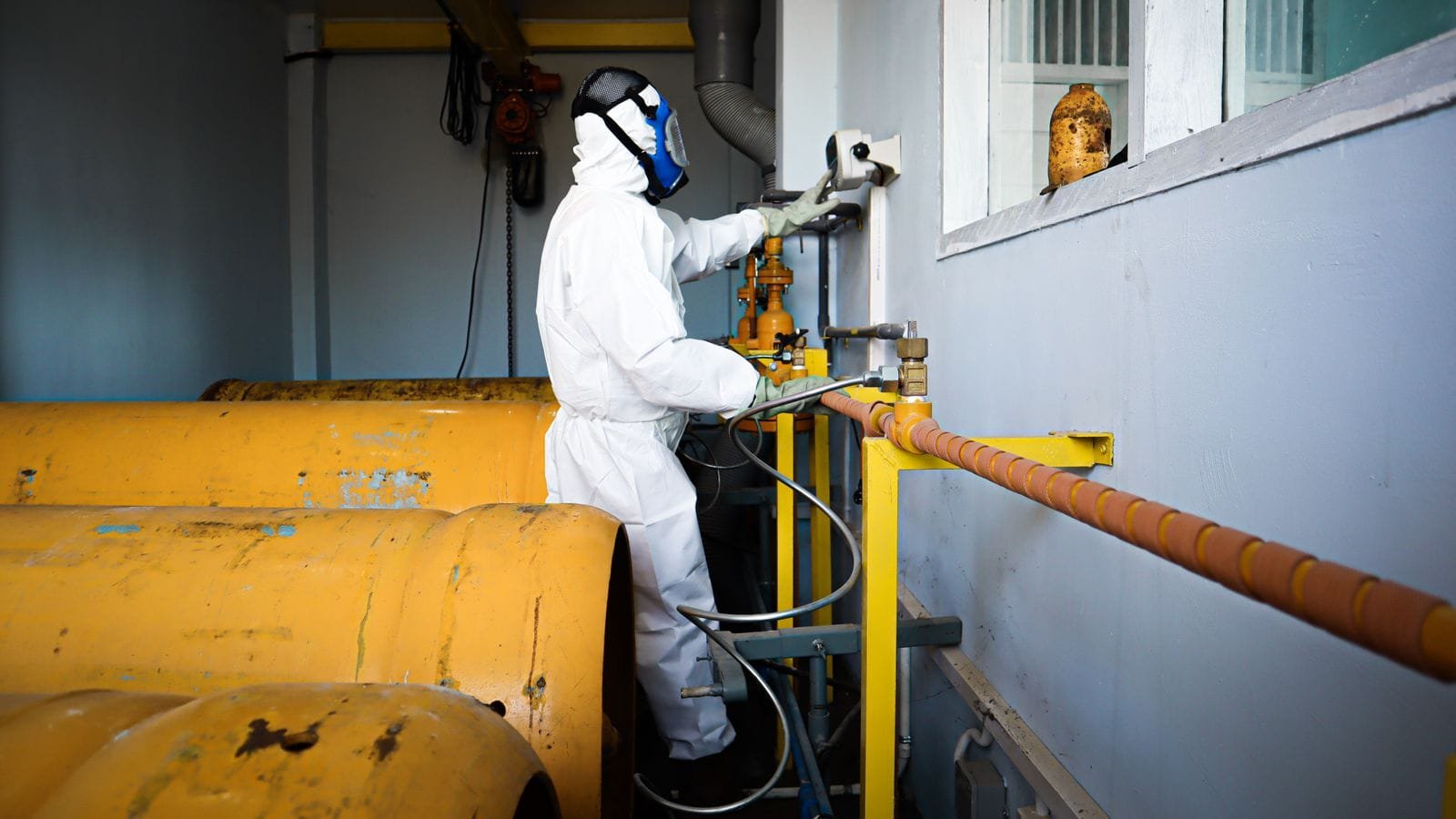 A worker in a hooded coverall, face shield and respirator opens a valve on chemical pipework beside large yellow storage tanks. Photo: Govin MU, via Pexels