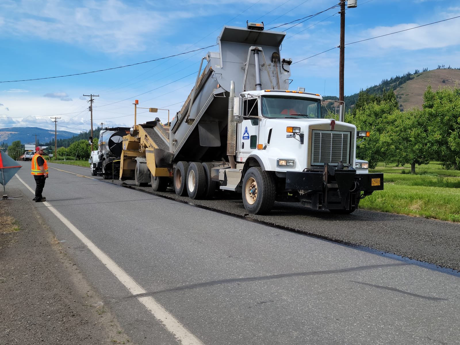 A highway paving crew at work: a dump truck backing to a paver while a worker in a high-visibility vest stands on the shoulder a few feet from the moving equipment. Photo: Tom Shamberger, via Pexels (Pexels Licence)