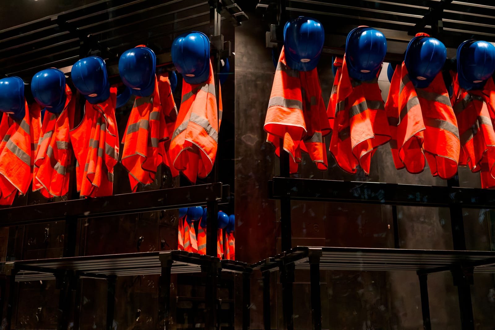 Rows of blue hard hats hanging above orange high-visibility vests on changing-room racks. Photo: Carsten Ruthemann, via Pexels (Pexels Licence)