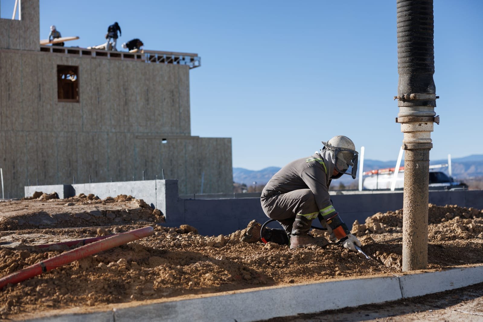 A construction worker in a hard hat and face shield kneels in the dirt beside a concrete line while a framing crew works on the roof deck behind. Photo: Rachel Tolander, via Pexels