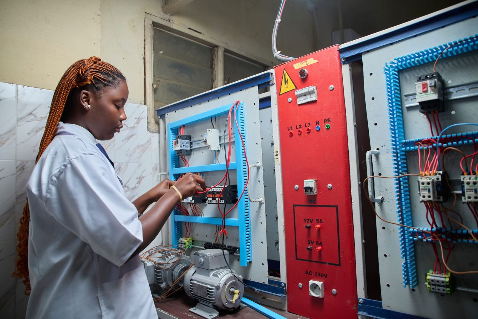 A technician works inside an open electrical control cabinet, wiring breakers and contactors mounted on rails. Photo: mickael ange konan, via Pexels
