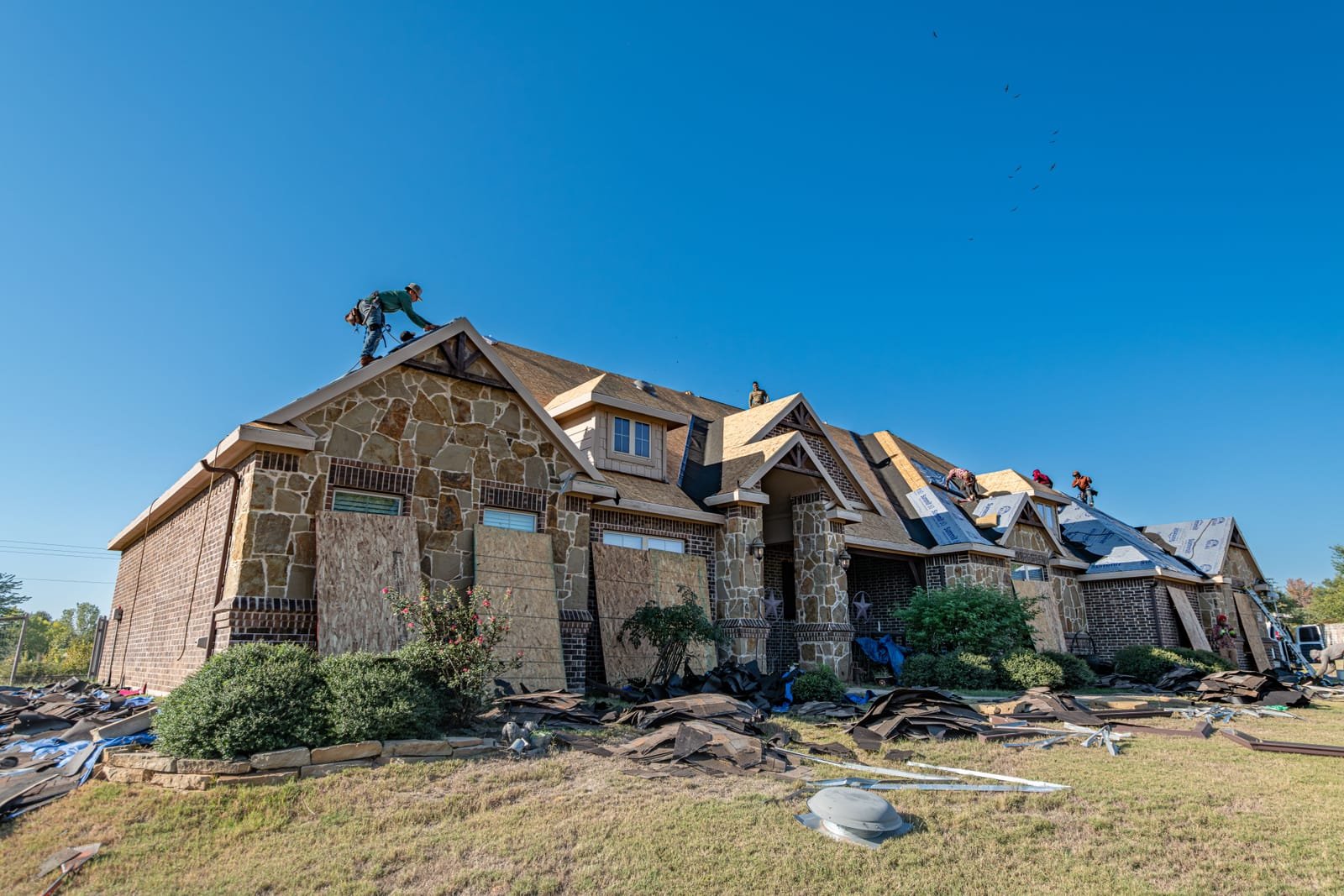 Roofers working along the ridge and slope of a brick house during a residential roof replacement under a clear sky. Photo: Ryan Stephens, via Pexels