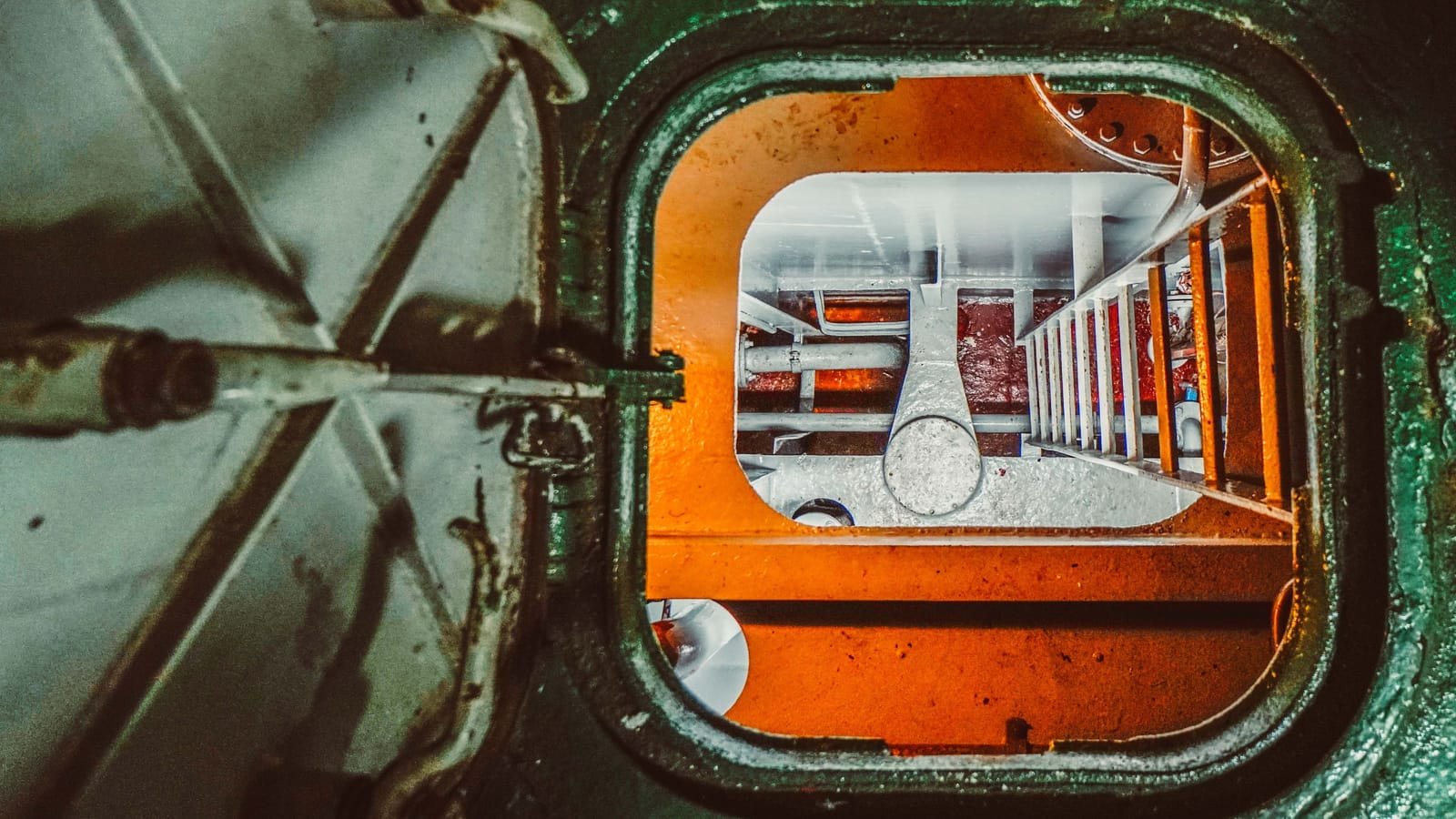 An open hatch cover on a steel deck, looking down a fixed ladder into an orange-lit compartment of pipework below. Photo: Nata Romualdo, via Pexels